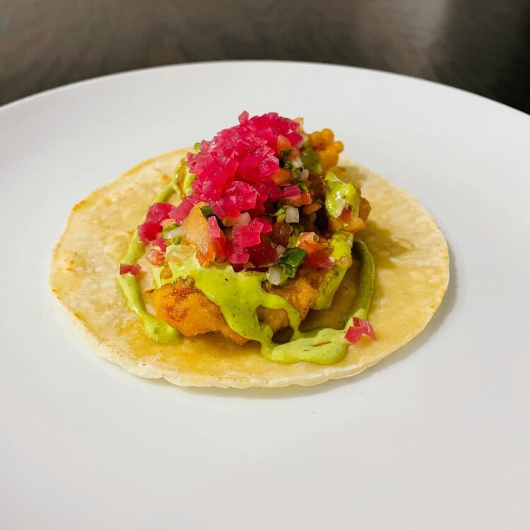 Fish taco topped with pico de gallo and pickled red onion on white plate