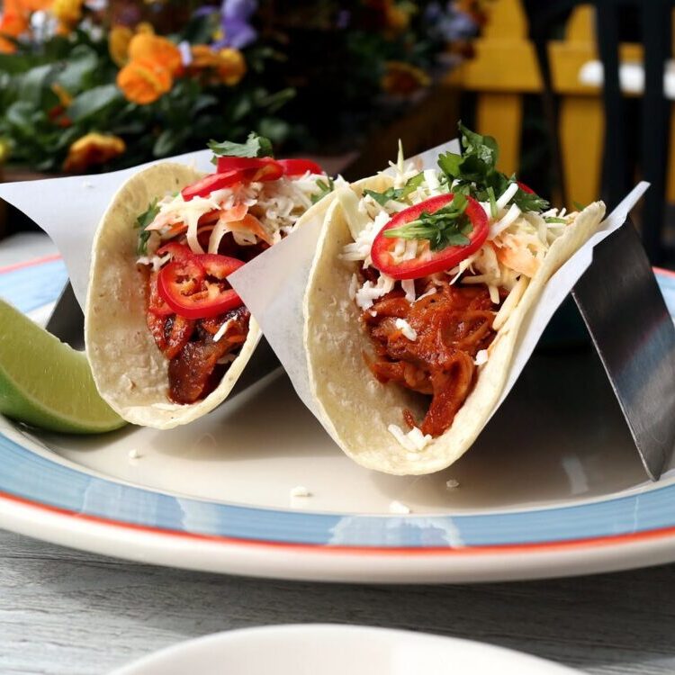 Two soft tacos filled with braised meat, shredded cheese, red pepper slices, and cilantro on a blue-rimmed plate with a lime wedge