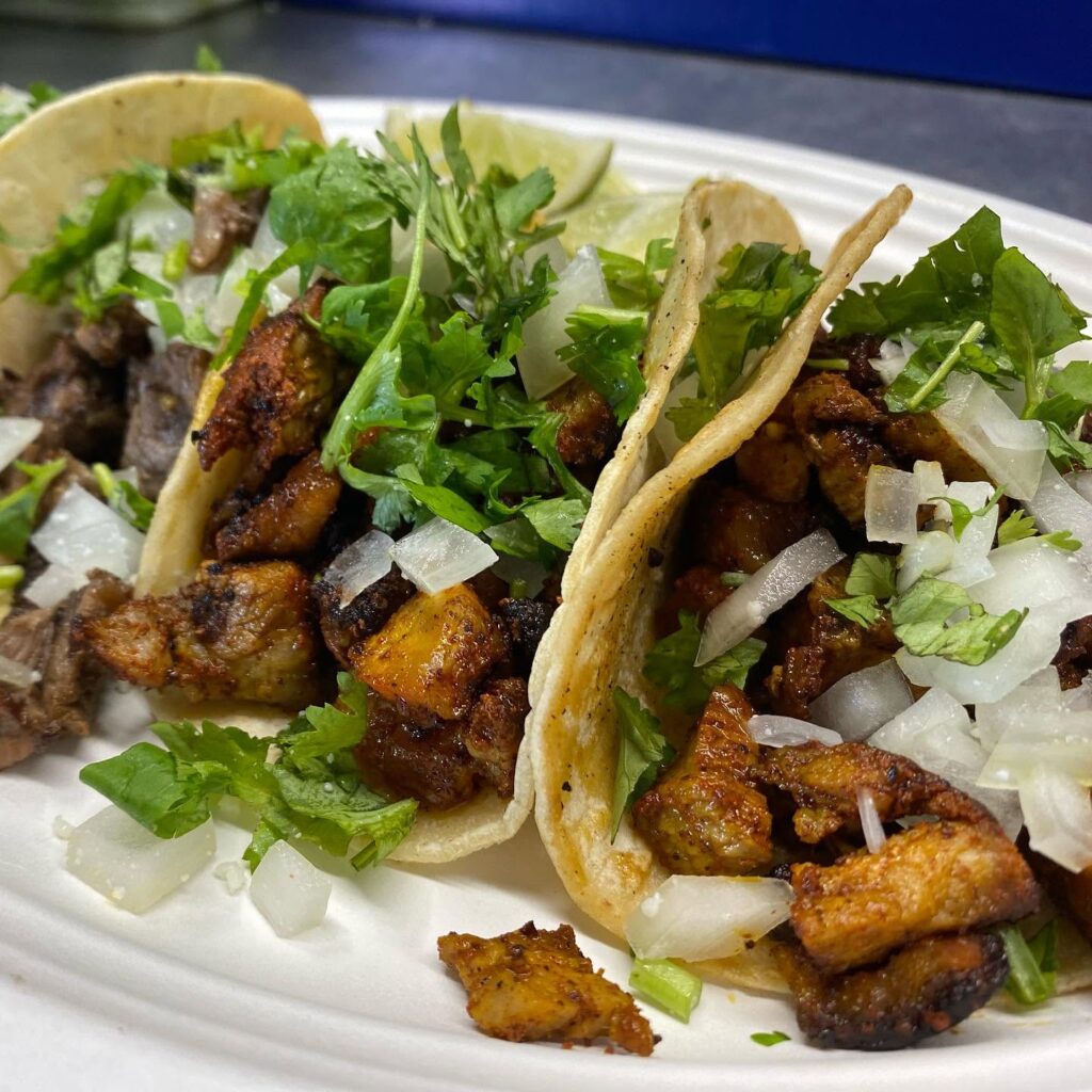 Two soft corn tortillas filled with seasoned grilled steak, chopped onions, and fresh cilantro on a white plate, with lime wedges nearby
