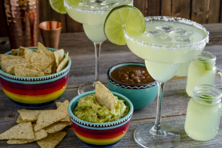 Three margarita glasses with salted rims and lime wedges, surrounded by bowls of guacamole and salsa with tortilla chips on a wooden table.