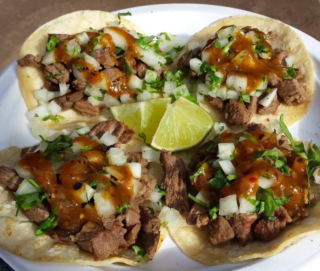 Four beef tacos on small corn tortillas topped with diced onions, cilantro, hot salsa, and lime wedges on a white plate.
