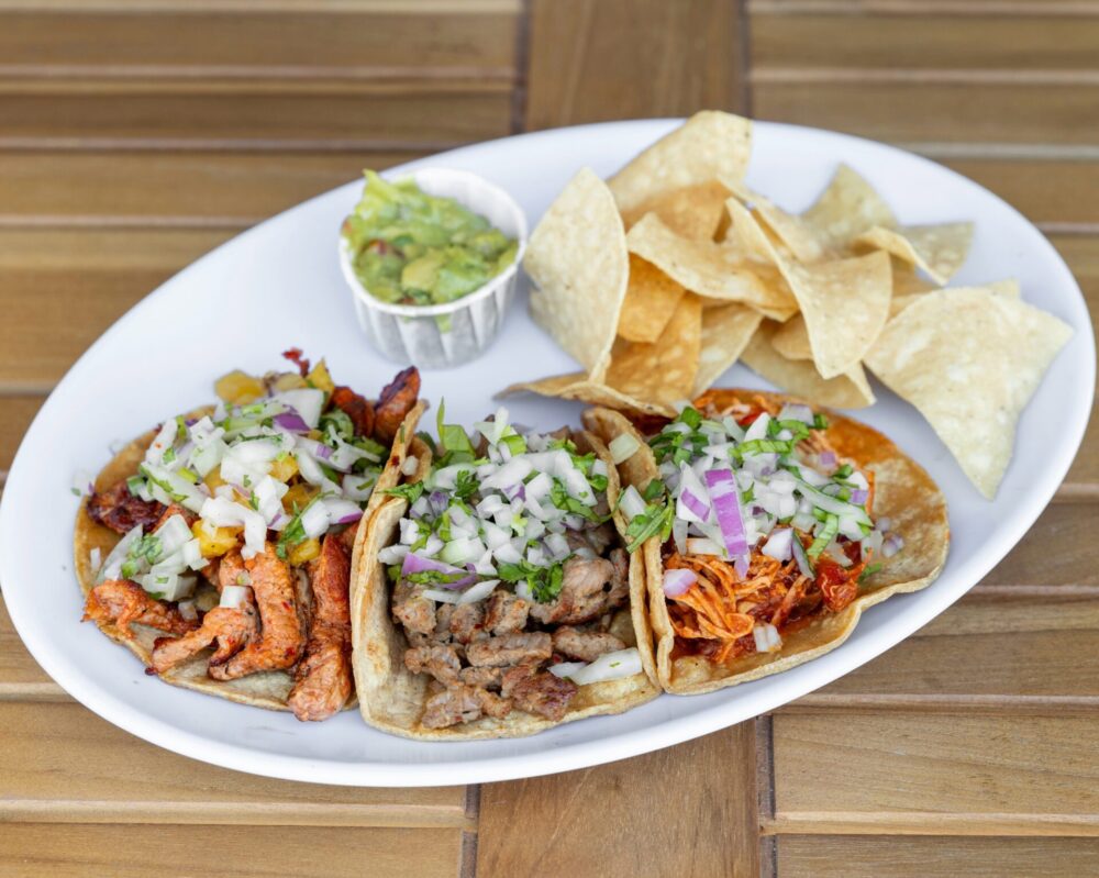 Plate with several tacos topped with diced onions and cilantro, surrounded by tortilla chips and a small cup of guacamole and salsa on the side.