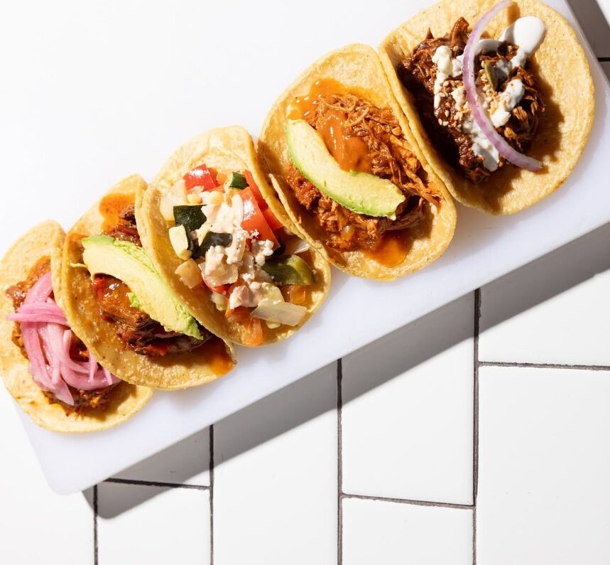 Five soft corn tacos arranged on a white rectangular platter, each with different fillings like pulled pork, avocado, and onions.
