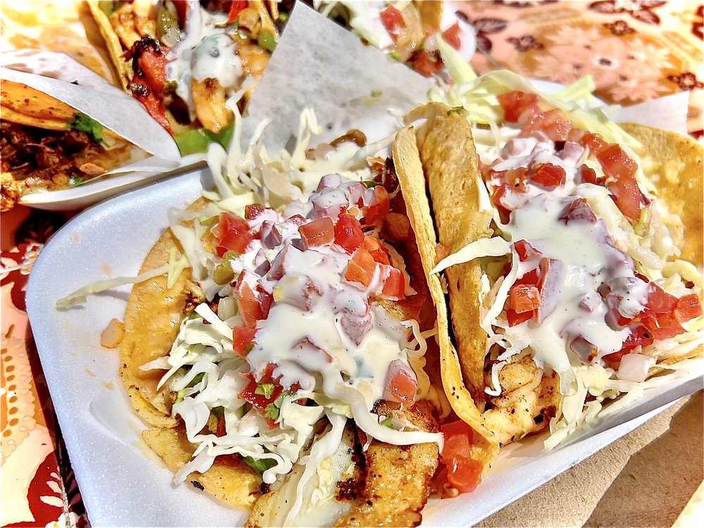 Two tacos filled with seasoned meat, shredded cabbage, diced tomatoes, and creamy sauce on a foam plate with tortilla chips nearby.
