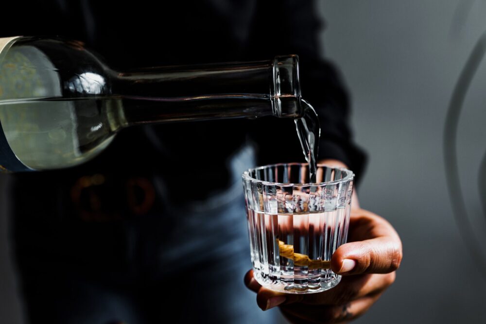 Close-up of a hand pouring a clear drink from a bottle into a crystal glass against a dark background.