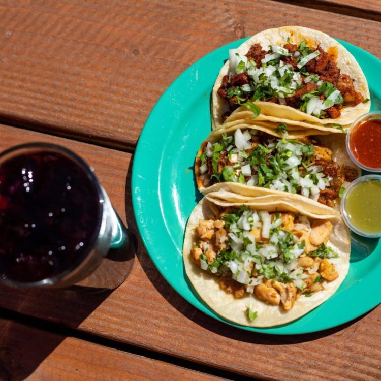 Three tacos topped with onions and cilantro on a turquoise plate, with red and green salsas and a glass of red beverage on a wooden table.