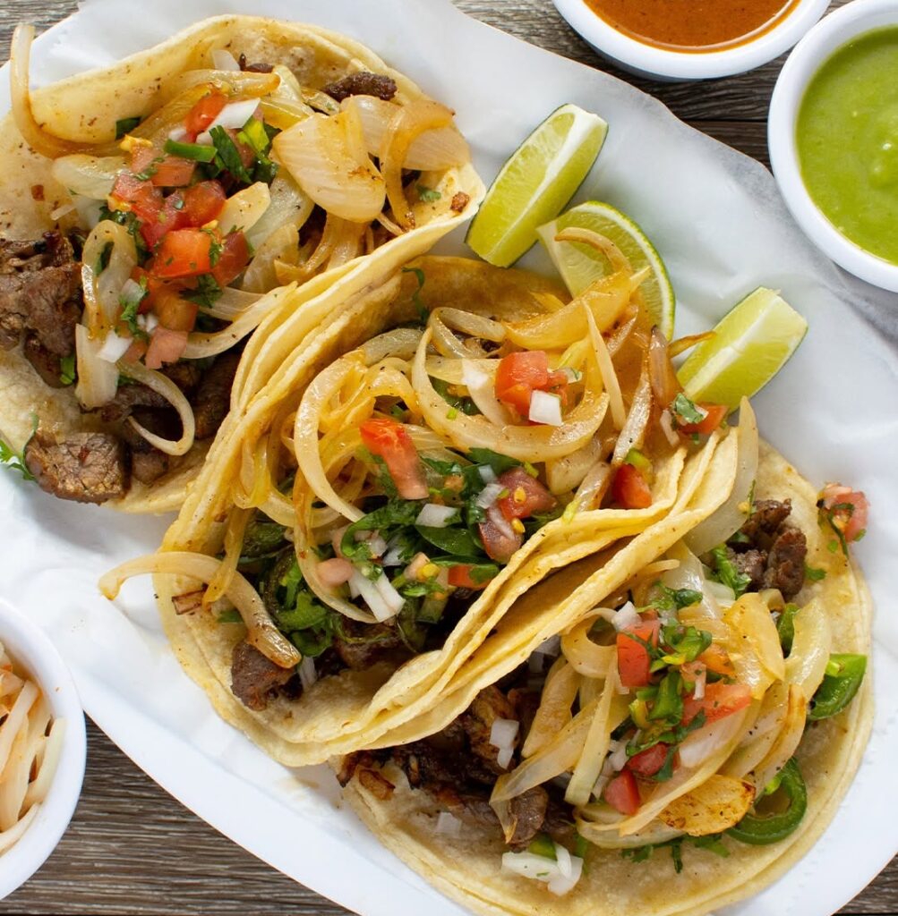 Two beef street tacos filled with onions, cilantro, and tomatoes on warm tortillas, with lime wedges and sauces on the side.