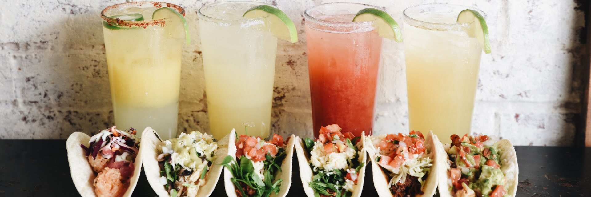 Five colorful cocktails with lime wedges behind a row of tacos on a dark table