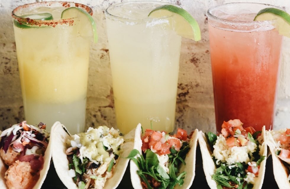 Five colorful cocktails with lime wedges behind a row of tacos on a dark table