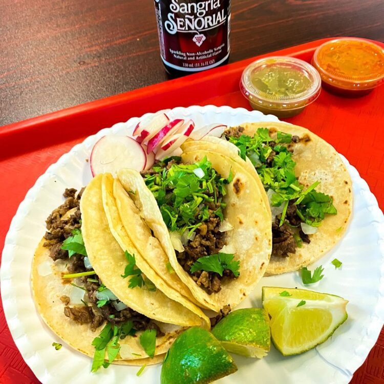 Tacos with beef, chopped cilantro and onions on soft tortillas, lime wedges and radish slices on the plate; sauces in the background.