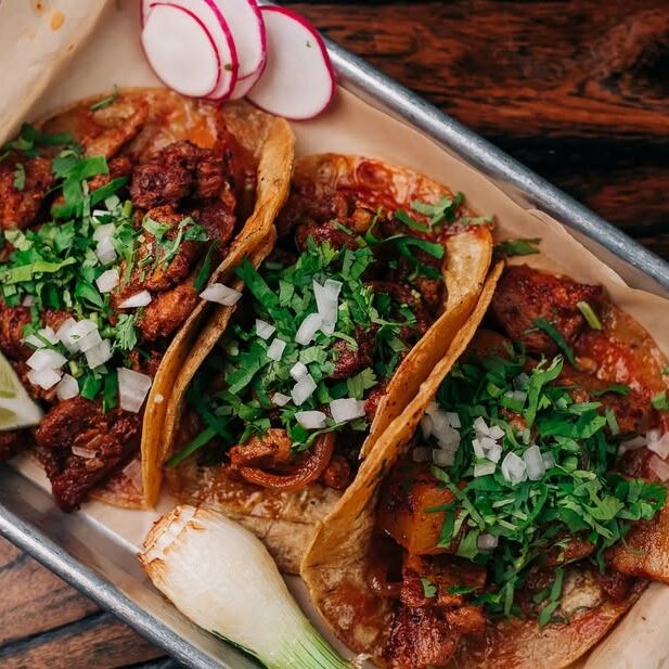 Three tacos filled with seasoned meat, topped with onions and cilantro, on a parchment-lined tray with lime wedges and radish slices.