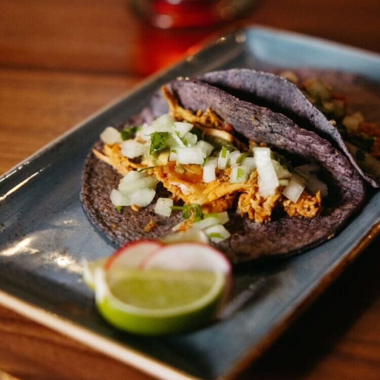 Two blue corn tortillas filled with shredded chicken, diced onions, and cilantro on a plate with a lime wedge.