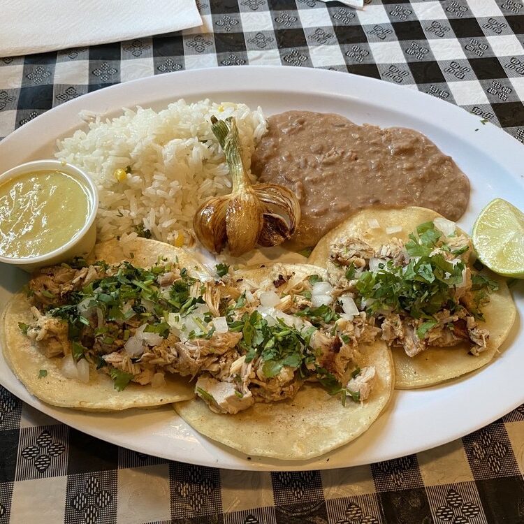 Plate of fish/taco-style tacos with shredded chicken, onions, and cilantro, served with white rice, refried beans, a lime wedge, roasted garlic, and a side sauce in a cup.