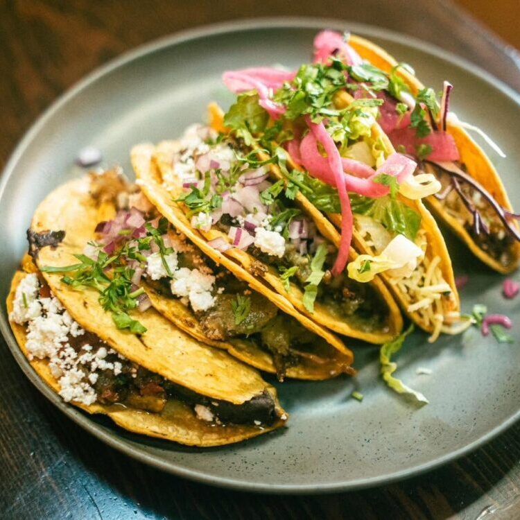 Three beef tacos on a gray plate, topped with onions, cilantro, crumbled cheese, and pickled red onions.