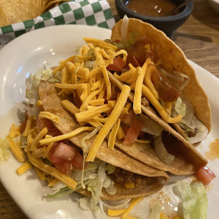 Two hard-shell tacos filled with lettuce, diced tomatoes, seasoned meat, and shredded cheddar on a white plate with chips and salsa in the background