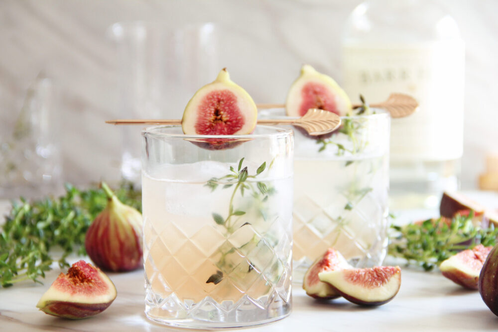 Cocktails in crystal glasses garnished with fig halves and thyme, with sliced figs on a marble surface in the background.