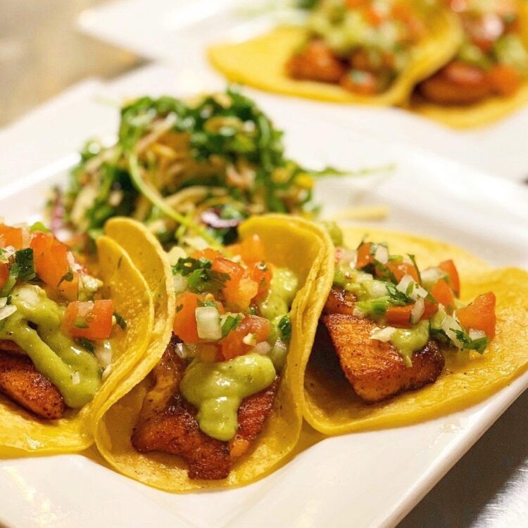 Three tacos on a white plate with crispy tortillas, filled with seasoned meat, avocado crema, and pico de gallo.