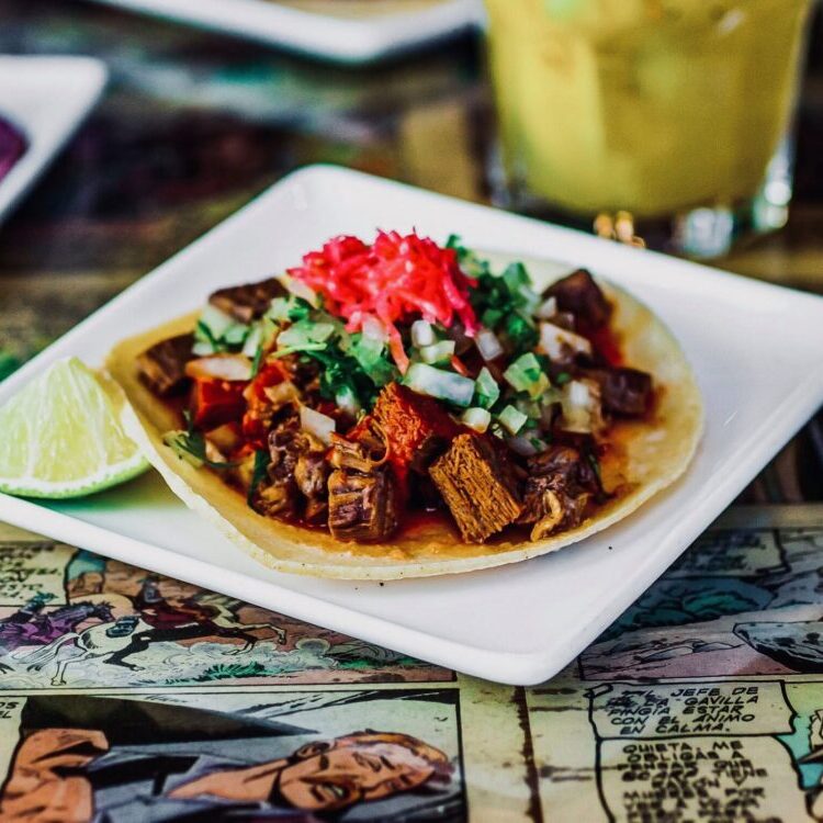 Taj&iacute;n-spiced tacos topped with diced onions, cilantro, and red pickled onions on a white plate with lime wedges