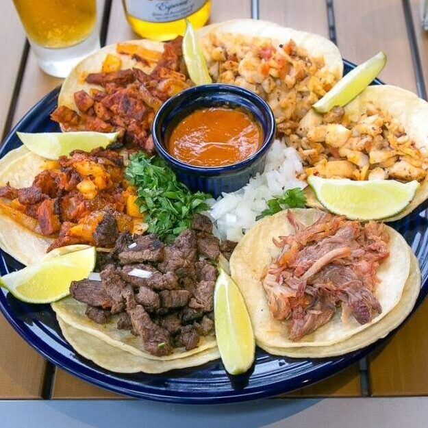 Plate of assorted tacos with beef, chicken, and vegetables, lime wedges, cilantro, and a central bowl of sauce on a blue platter.