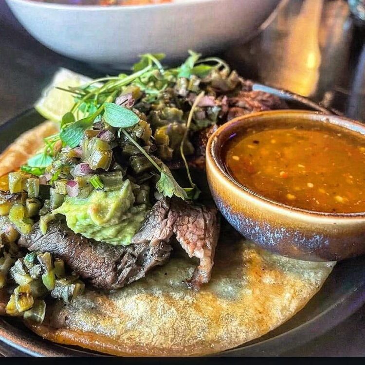 Beef steak on a warm tortilla, topped with guacamole, chopped onions and cilantro, with a side bowl of salsa.