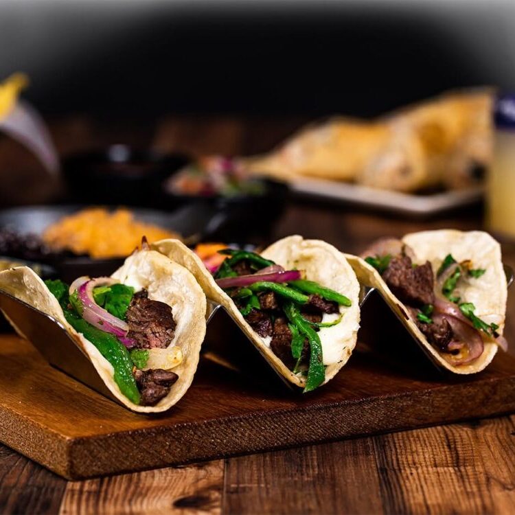 Three beef tacos with onions and green peppers on a wooden serving board, with chips and a drink in the background.
