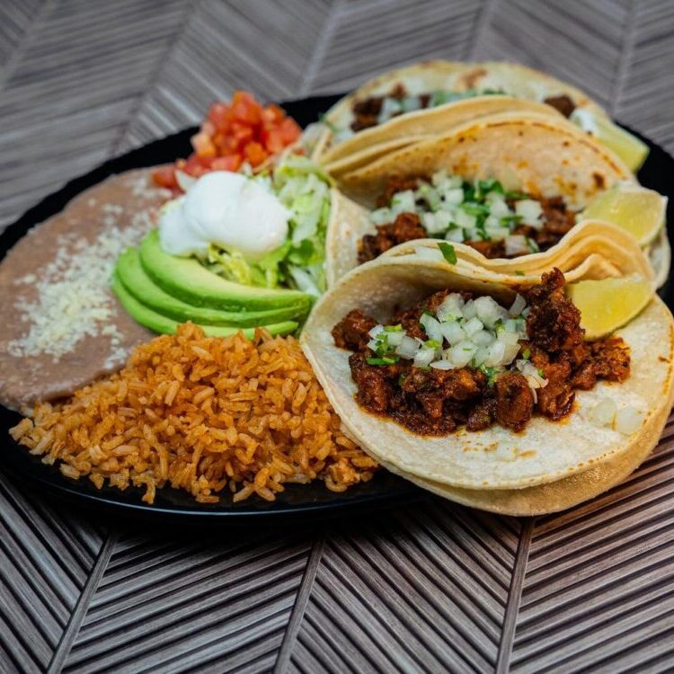 Three soft tacos filled with seasoned meat, topped with onions and cilantro, served with lime wedges and avocado slices on a black plate with rice, beans, and pico de gallo.