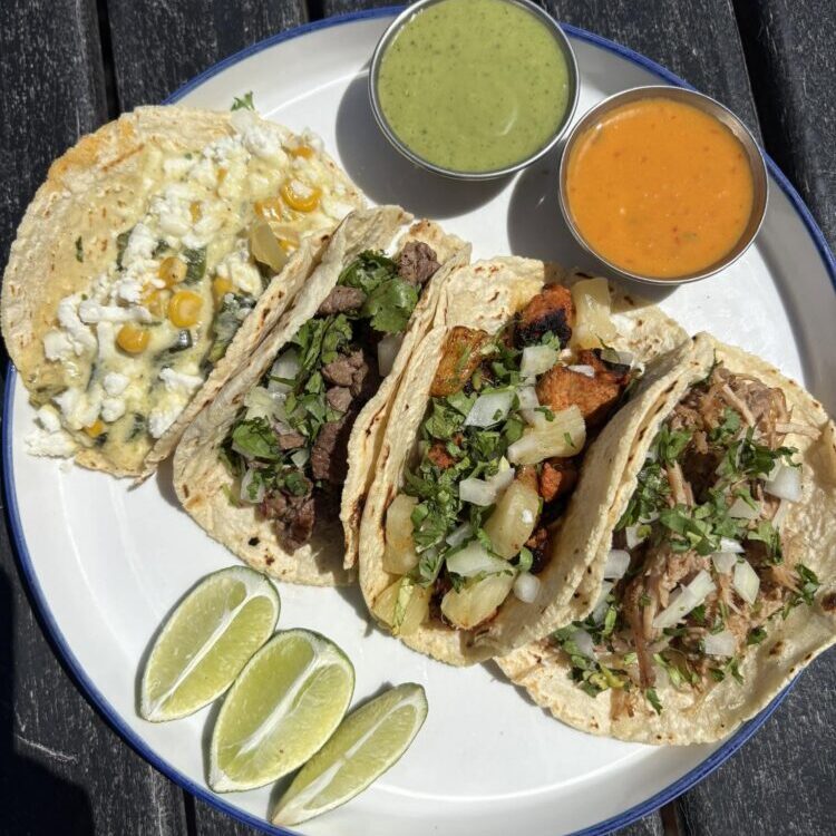 Plate of soft corn tacos filled with beef, onions, cilantro, and cheese, with lime wedges and two salsas (green and orange) on the side.