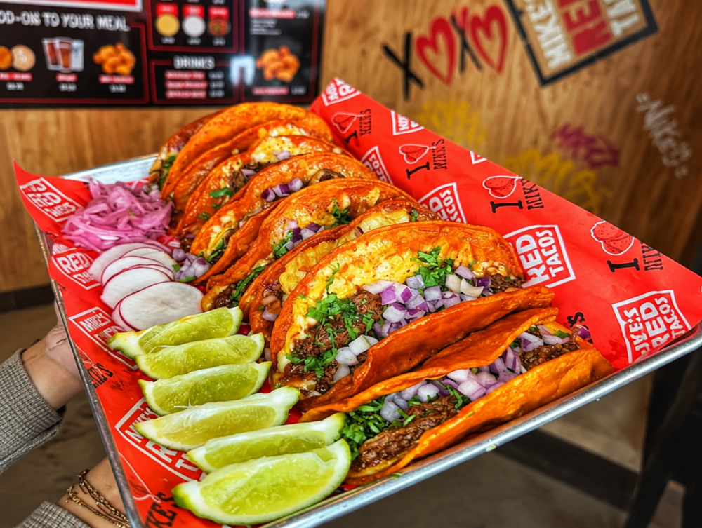 Tray of loaded beef tacos with cilantro and onions, lime wedges, radishes, and shredded red onions on a branded red liner.