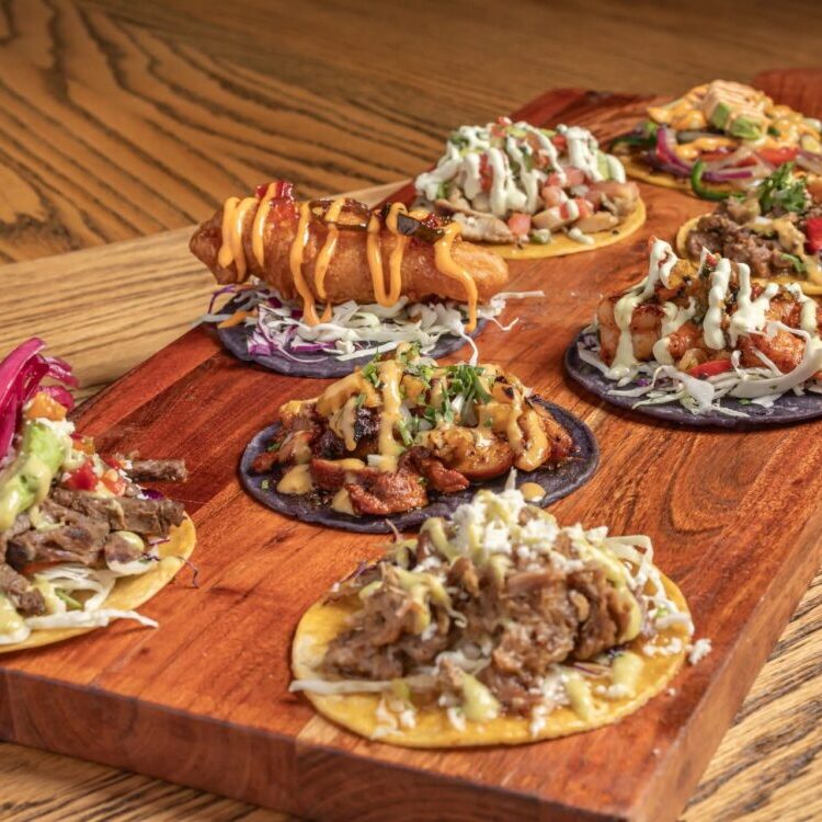 Assorted loaded mini tacos on a wooden serving board with colorful toppings and sauces.