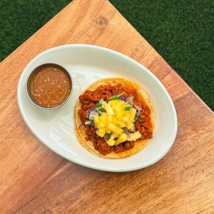 Taco on a white oval plate with chopped meat, pineapple salsa, onions and cilantro, served with a small cup of salsa on a wooden table outdoors.