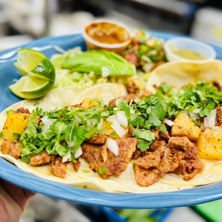 A blue plate piled with warm soft tacos, filled with tender seasoned meat, grilled pineapple, diced white onion, and fresh cilantro. Lime wedges, creamy avocado, salsa, and small cups of flavorful sauces complete this colorful Mexican mealserved fresh in Aurora for a bright, satisfying bite.