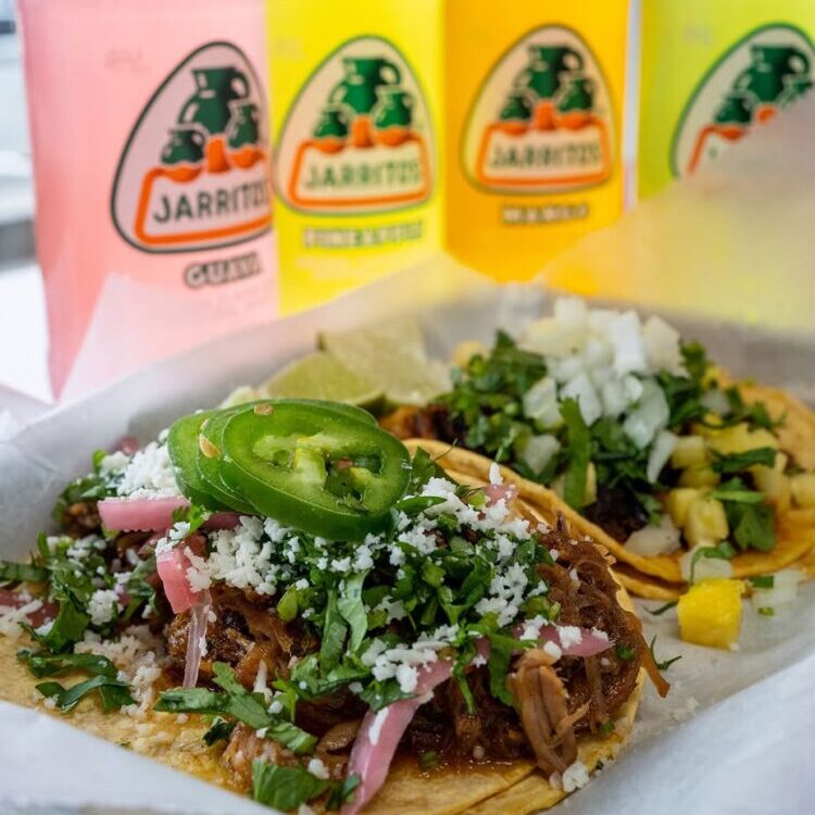 Tacos topped with cilantro, onions, cheese, and a jalape&ntilde;o slice, with colorful Jarritos bottles in the blurred background.