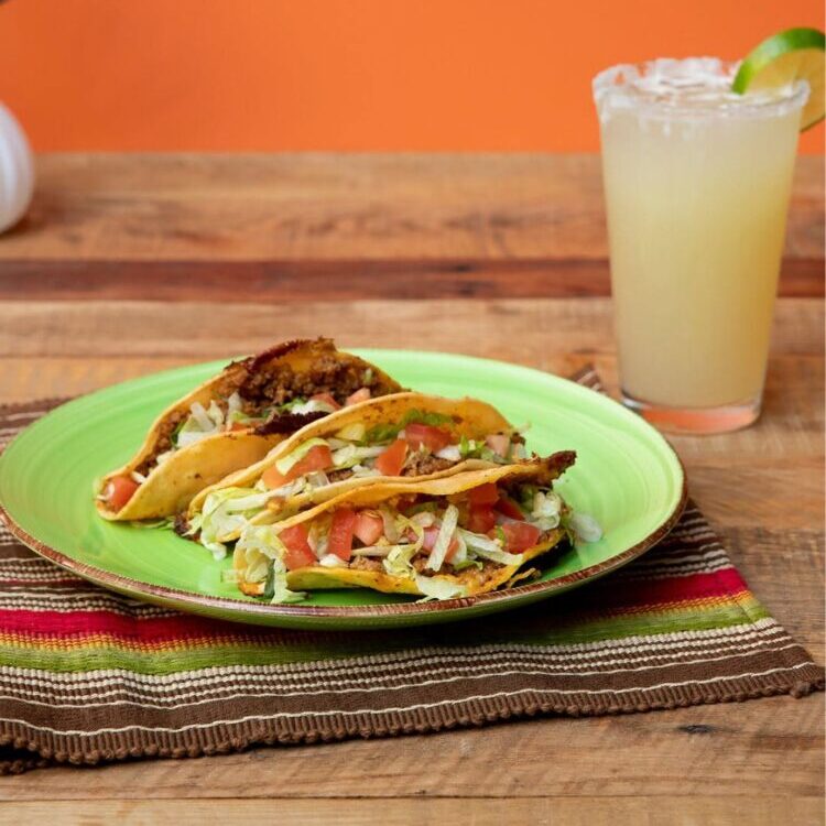 Two tacos filled with meat, lettuce, tomato on a bright green plate with a lime margarita beside them.