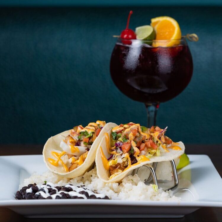 Taj&iacute;n-topped tacos filled with vegetables on a white plate, with rice and black beans nearby, in a close-up shot.