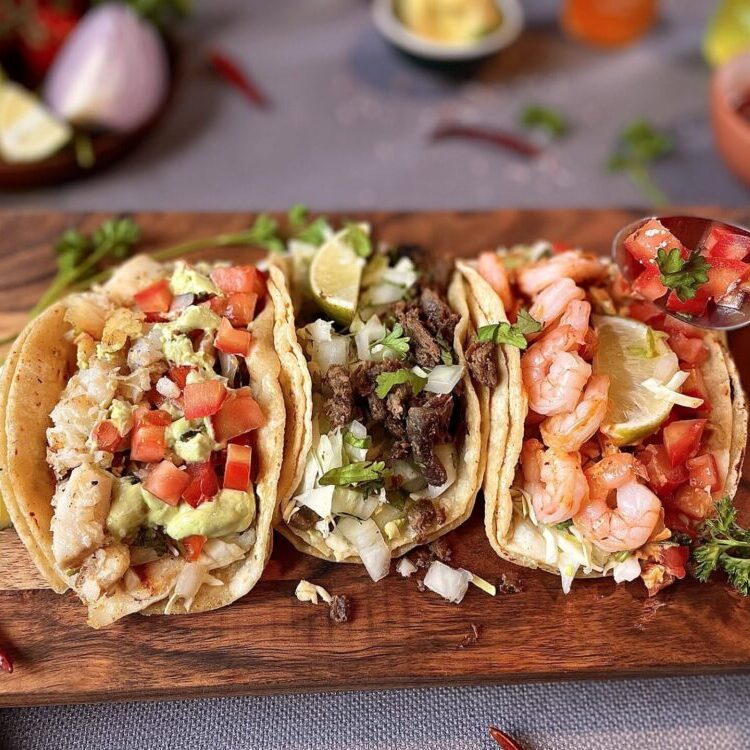 Tilled wooden board with three tacos filled with shrimp, beef, lettuce, pico de gallo, and lime wedges, garnished with herbs.