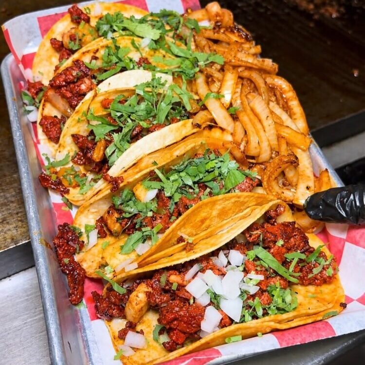 Tray of loaded street tacos on a metal tray with parchment, topped with chopped onions, cilantro, and red salsa sauce.