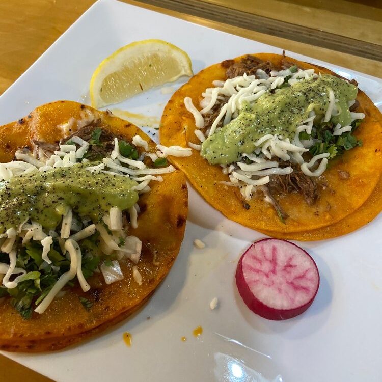 Two orange corn tortillas filled with shredded meat, onions, cilantro, and cheese, drizzled with green sauce on a white plate with a lemon wedge and a radish slice nearby.