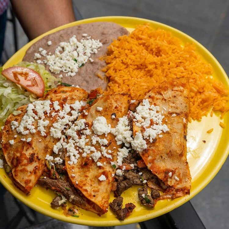 Grilled chicken fajitas with crumbled cheese on a yellow plate, served with rice, beans, lettuce, and tomato slices.